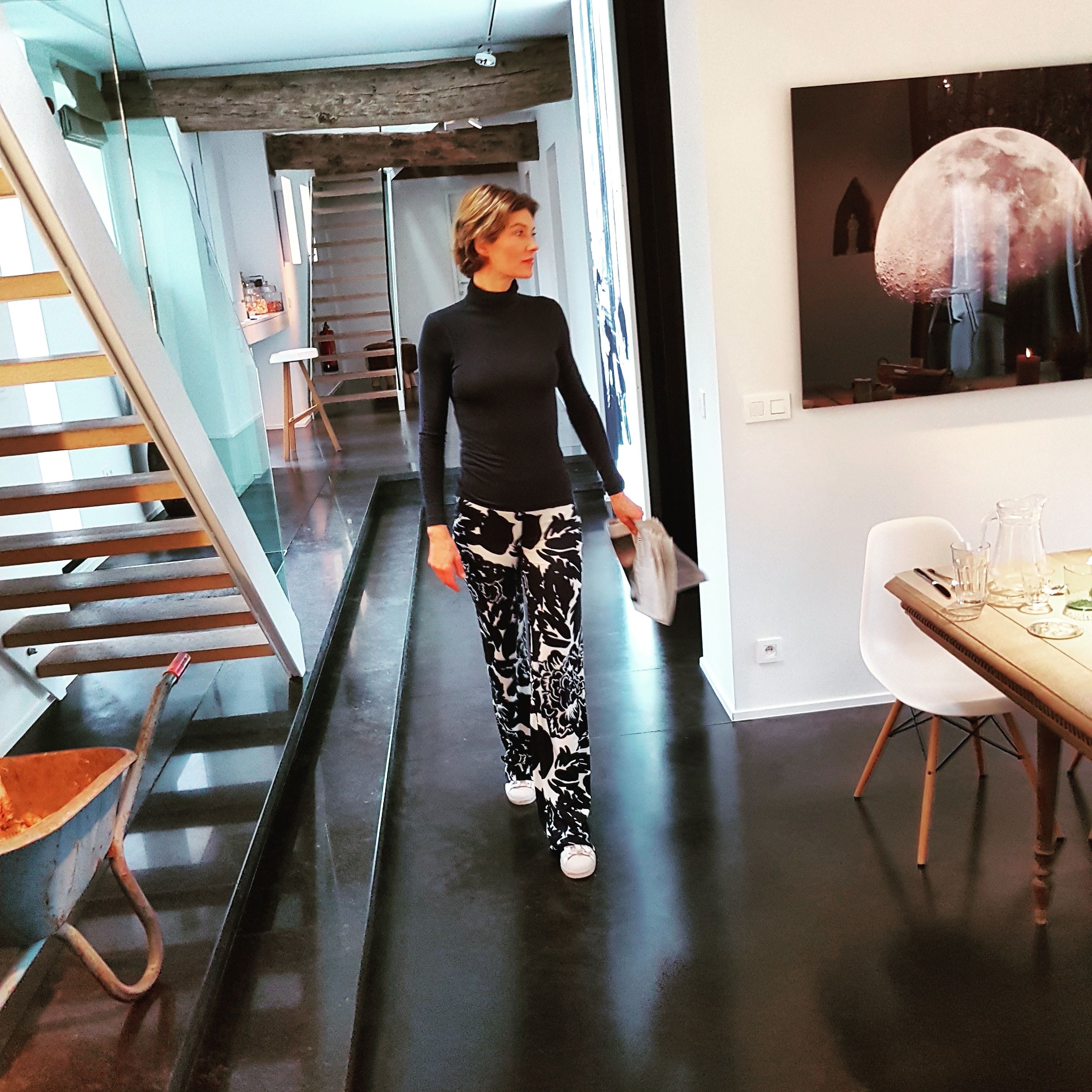 A woman walking through a modern interior with a staircase, a dining area, and a wall-mounted artwork.