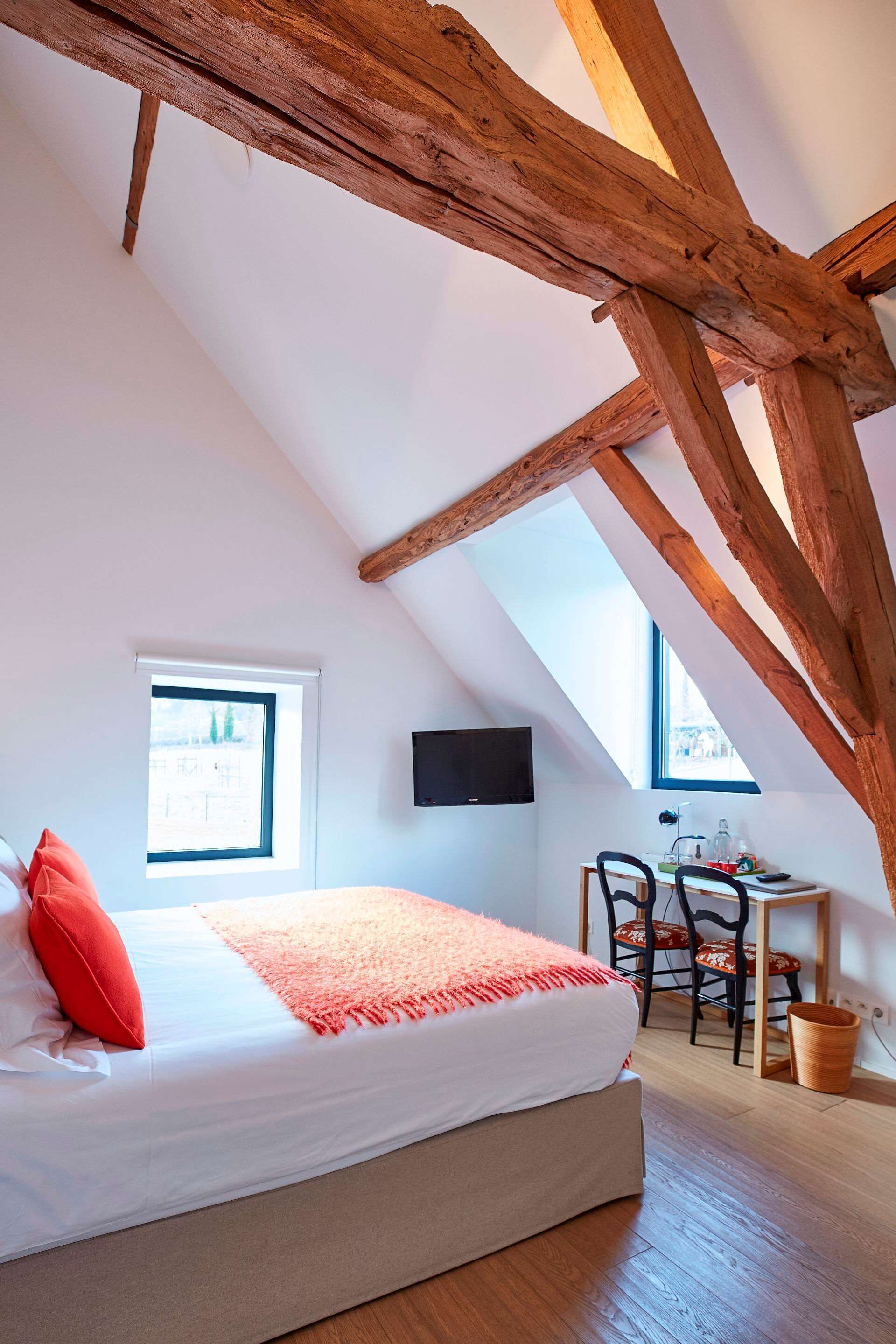 Un dormitorio con techo inclinado, una cama con almohada roja, un escritorio con sillas y un televisor en la pared.