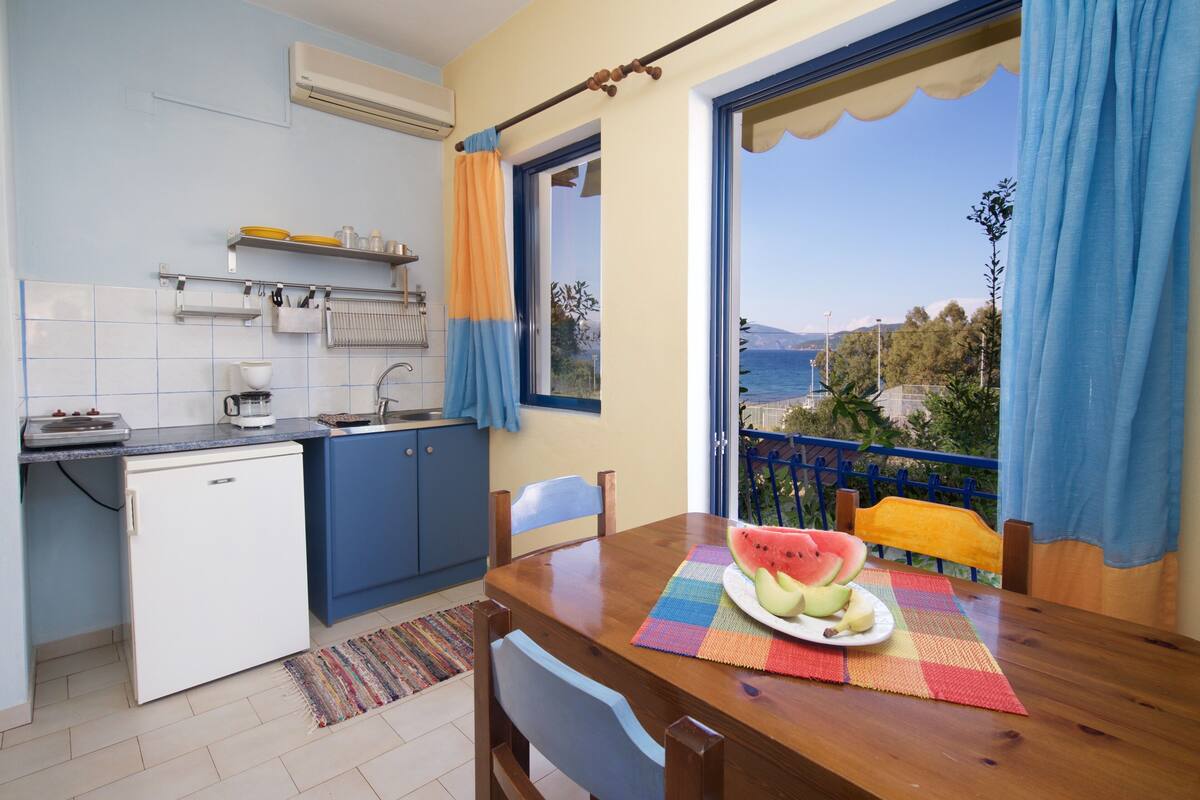 A compact kitchen with a dining area, a small refrigerator, and a view of the sea through a large window.