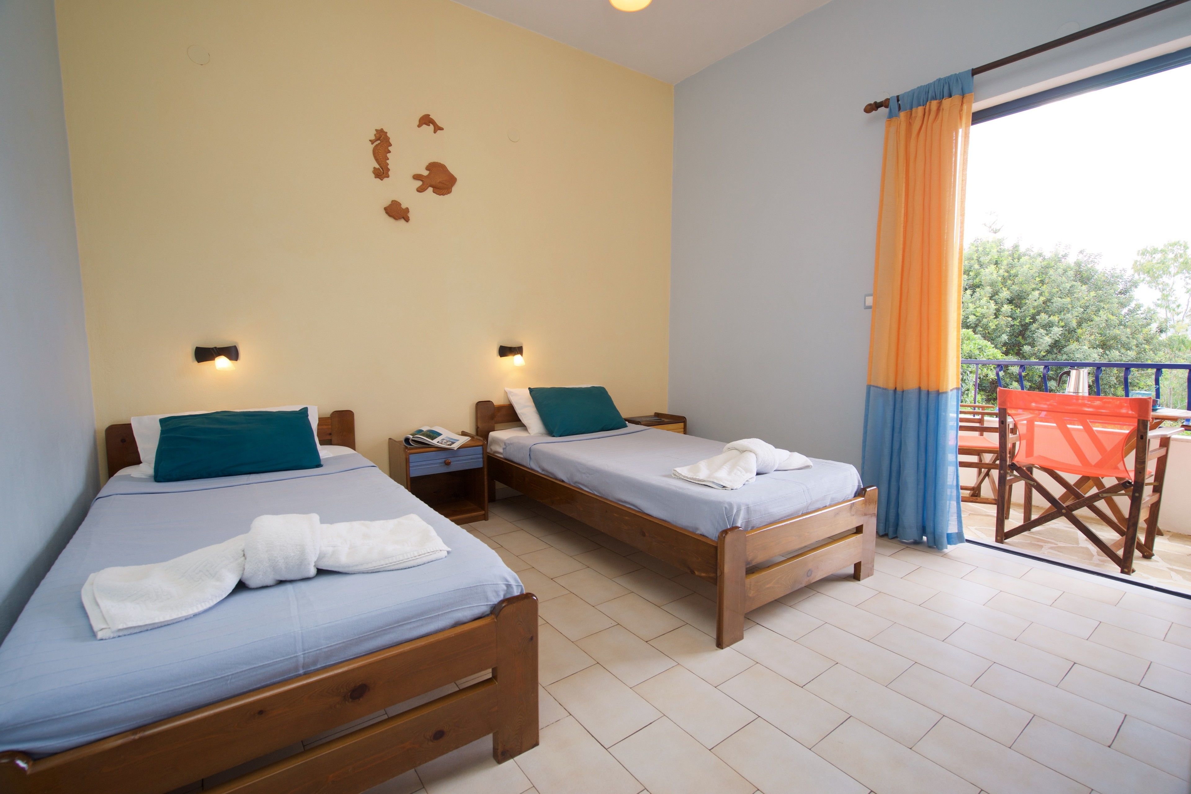 A room with two wooden beds, each with a blue mattress and a teal pillow. There are white towels on the beds. The room has a light-colored tiled floor and a window with blue curtains.