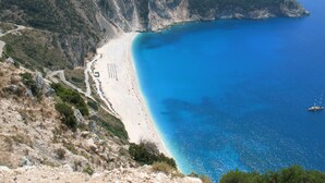 Una carretera costera que lleva a una playa con un promontorio rocoso y un mar azul transparente.