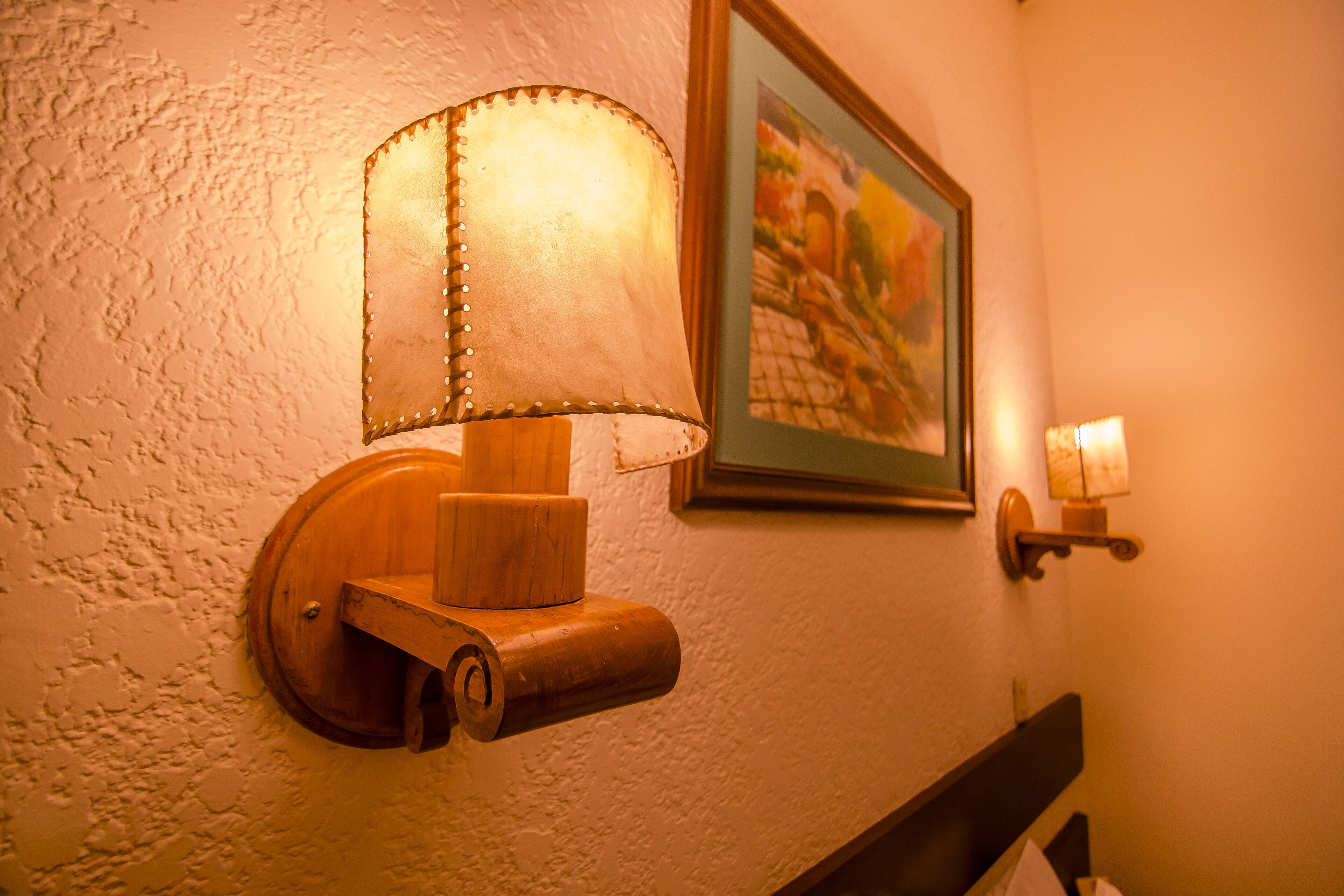 A wall-mounted lamp with a wooden base and a fabric shade, a framed picture above it, and a small table lamp to the right.