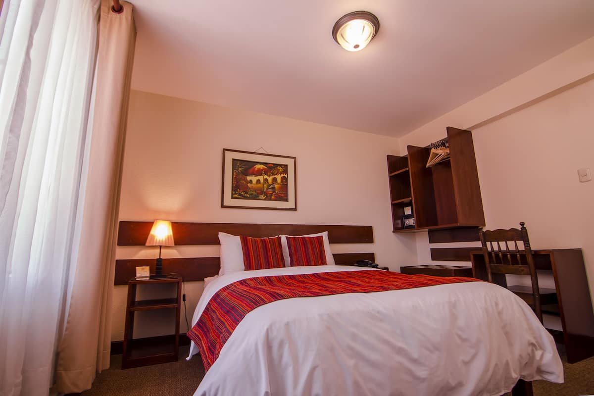 A hotel room with a neatly made bed, a bedside table with a lamp, a chair, a wardrobe, and a framed picture on the wall.