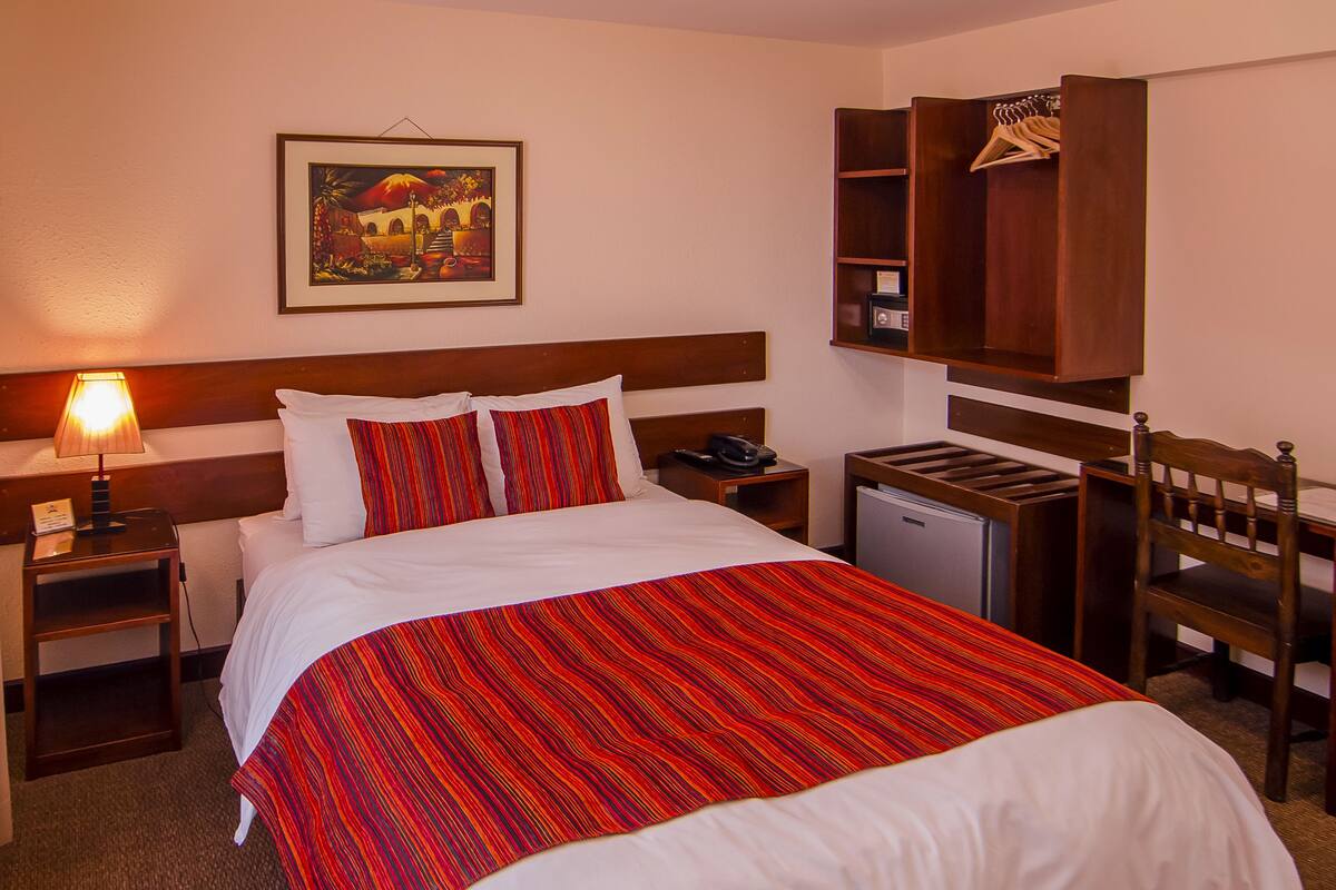 A hotel room with a bed, a nightstand, a desk, a chair, a wardrobe, a painting on the wall, and a lamp.