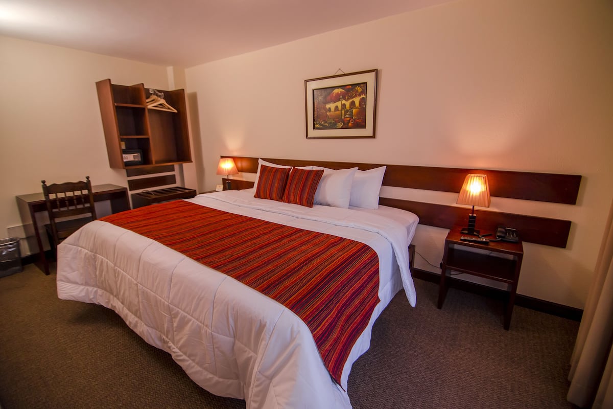 A hotel room with a large bed, a wooden headboard, a bedside table with a lamp, a desk with a chair, and a coat rack.