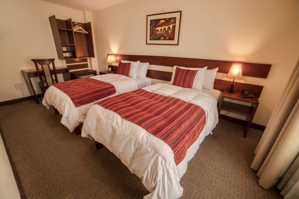 A hotel room with two beds, a wooden headboard, a nightstand with a lamp, a desk with a chair, and a coat rack.