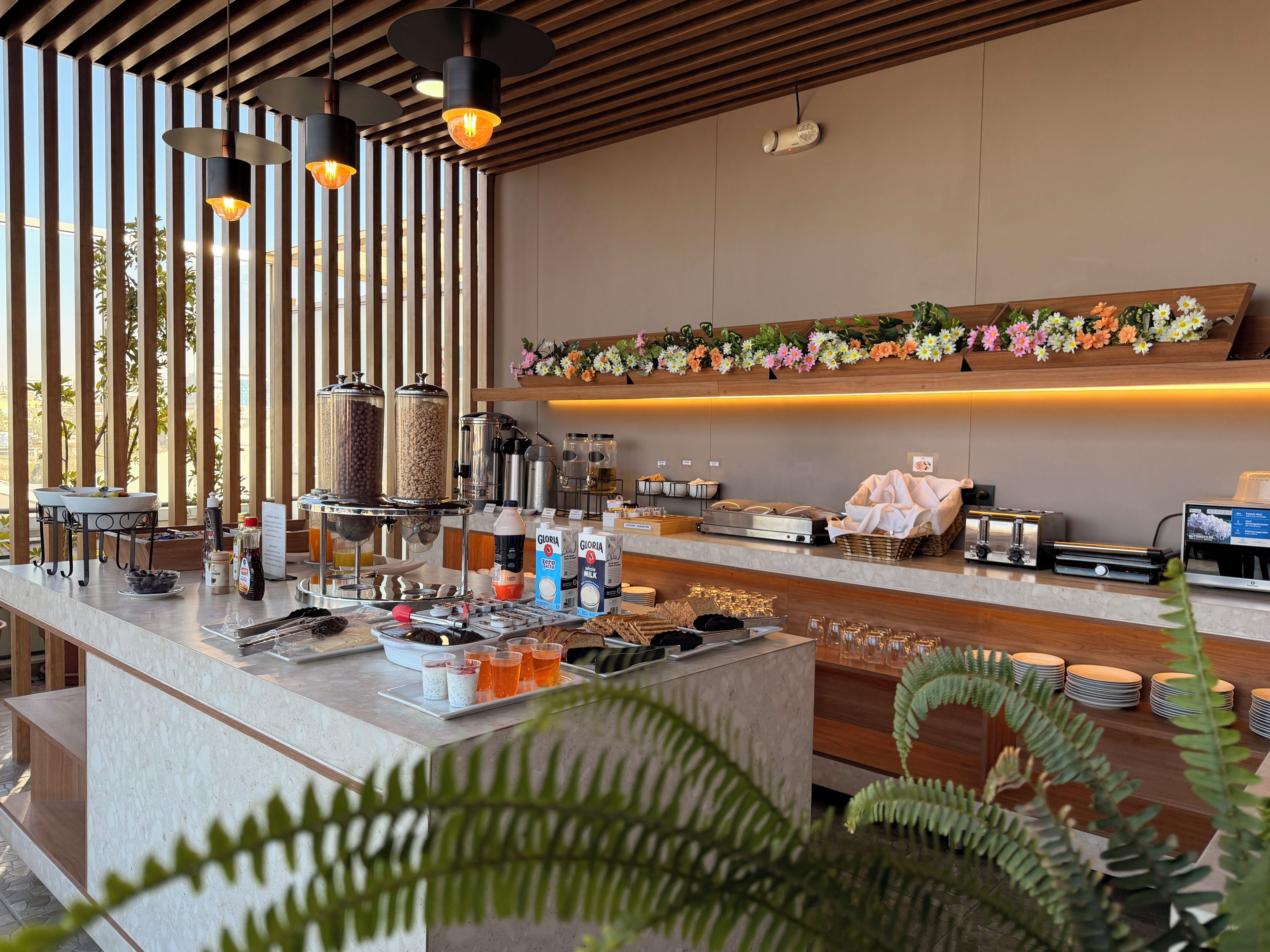 Una barra de desayuno moderna con máquinas de café, una presentación de frutas y un techo de madera.