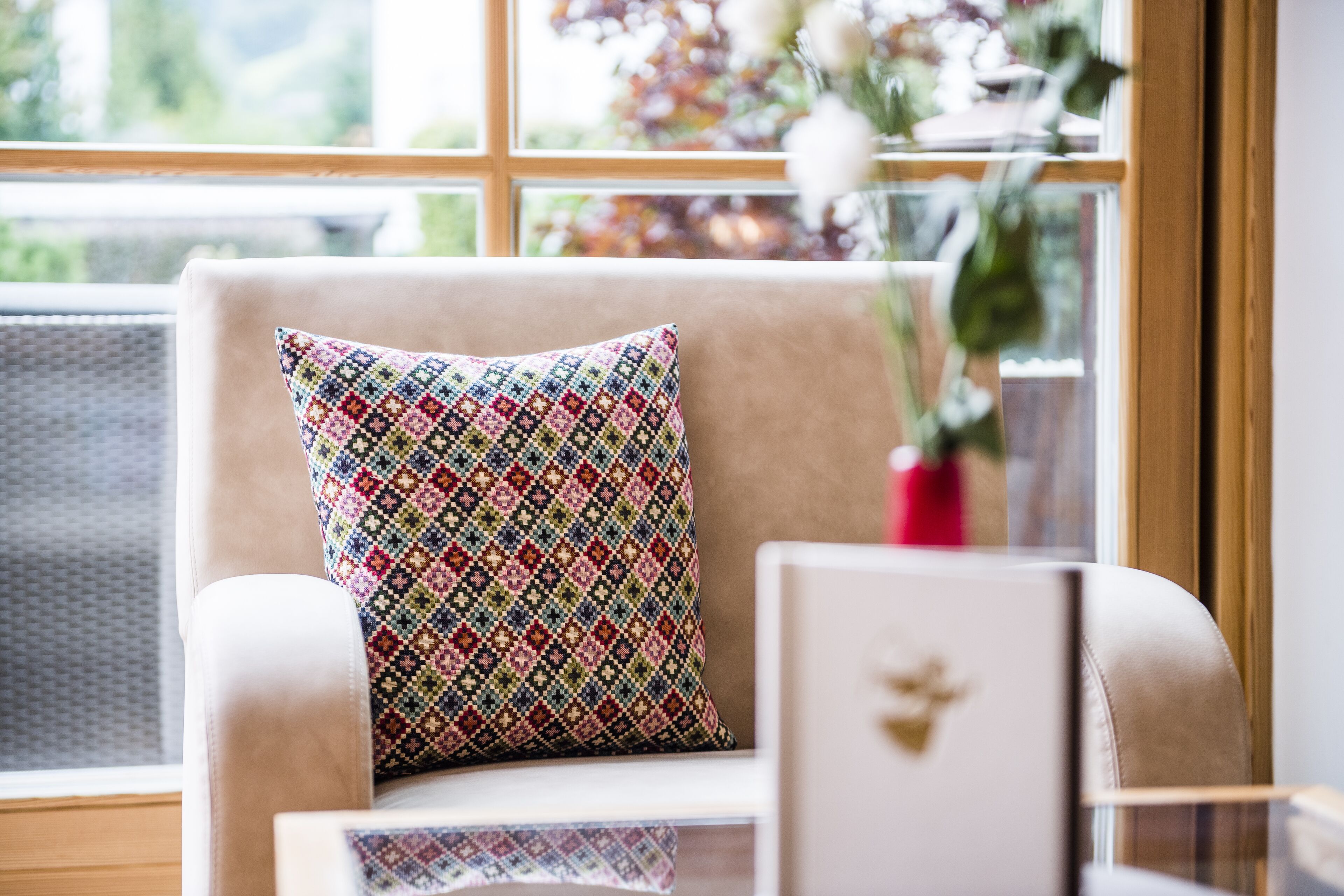 A chair with a patterned cushion and a vase with flowers on a windowsill.
