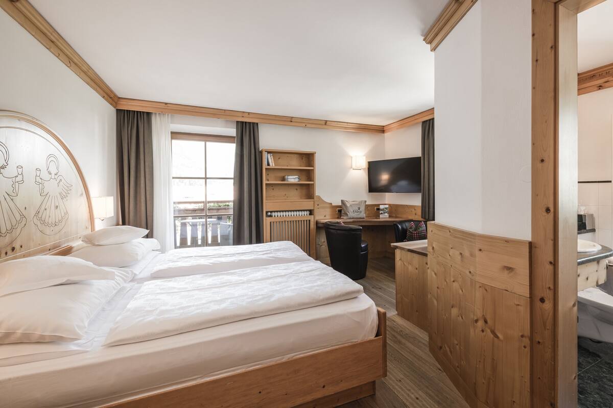 Una habitación de hotel con cabecera de madera, cama con ropa blanca, escritorio con silla, televisor y baño con inodoro y lavamanos.