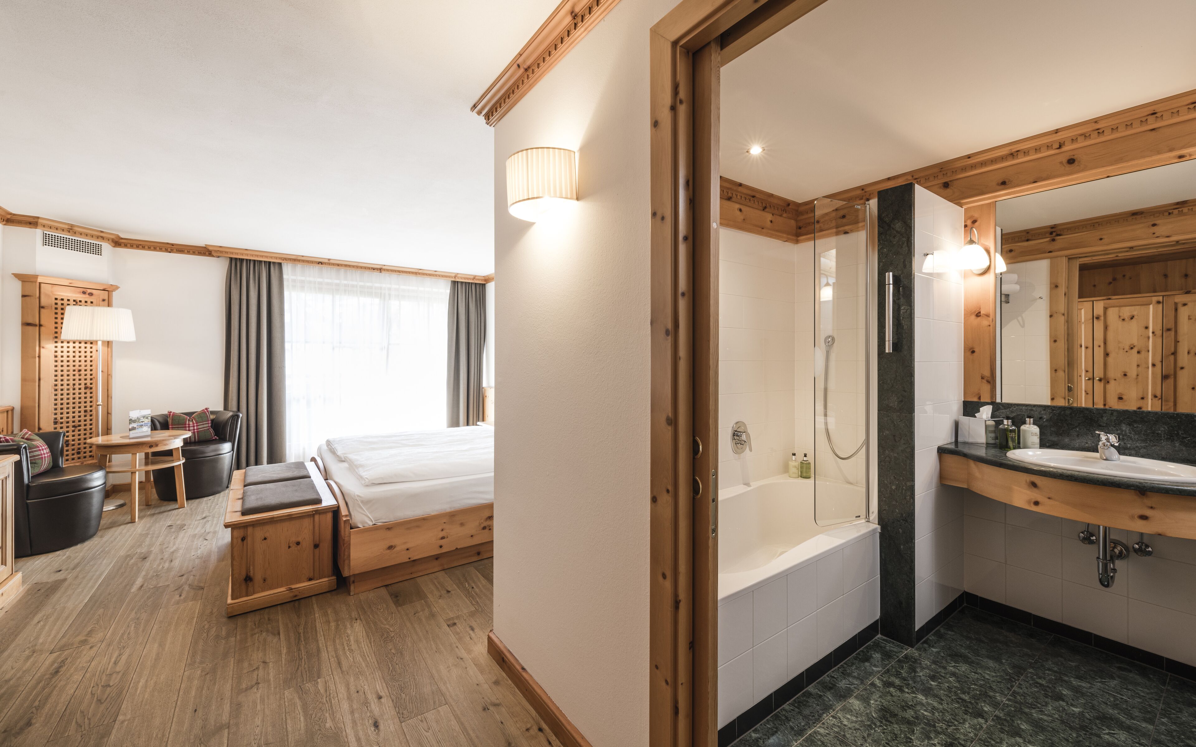 Ein Hotelzimmer mit Bett, einer Sitzecke mit Tisch und Stühlen, einem Badezimmer mit Badewanne und Waschbecken sowie Holzvertäfelung.