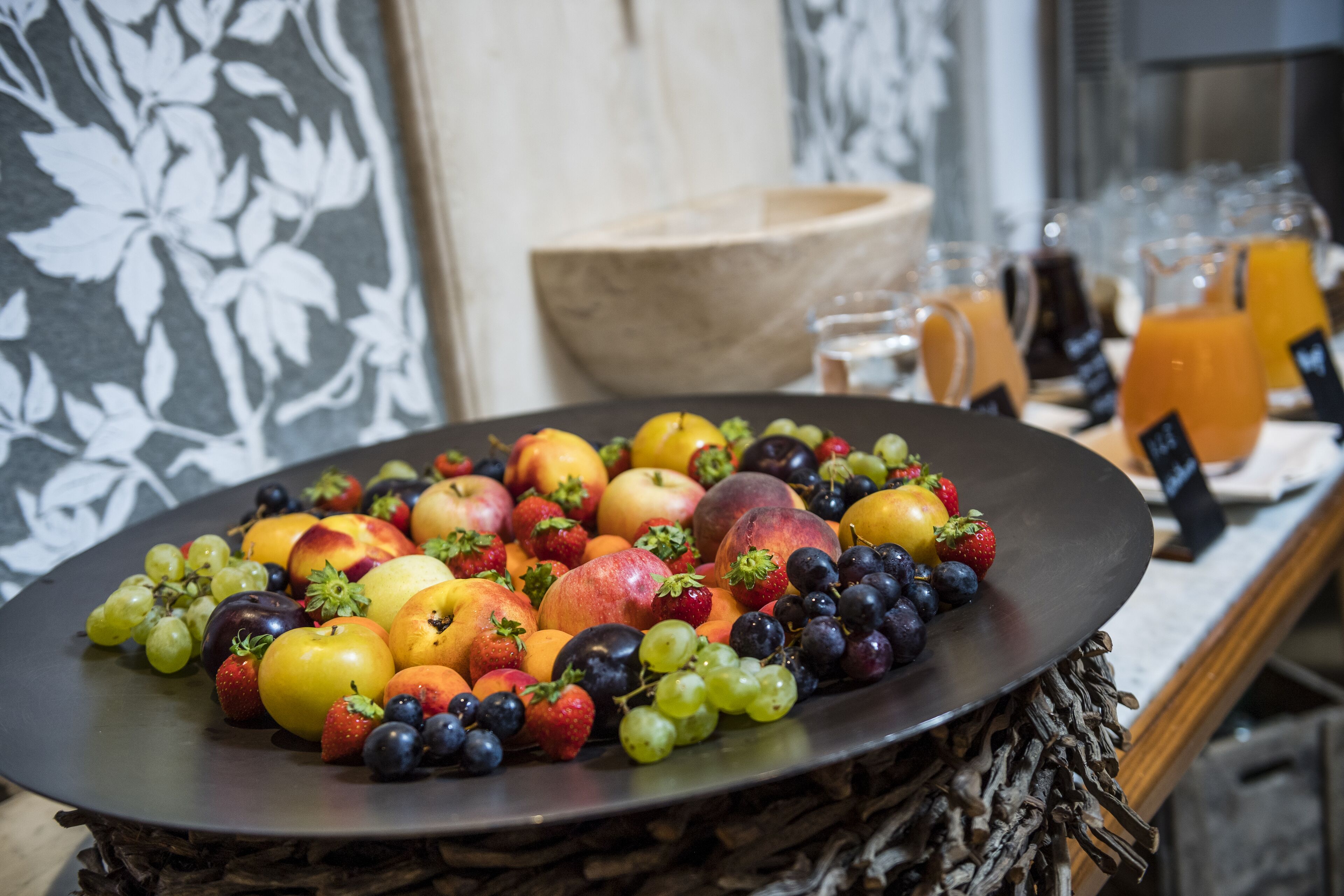 A fruit platter on a round table with a woven base, surrounded by a wooden bowl and various glass containers.