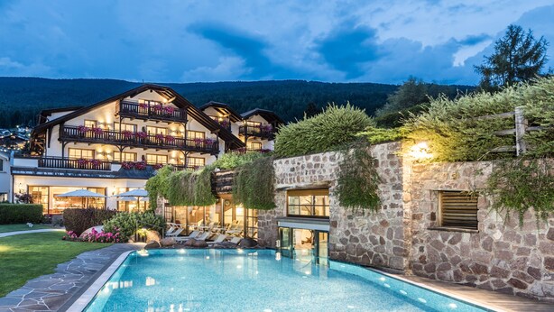 Un edificio de dos pisos con piscina, muros de piedra y balcones con plantas.