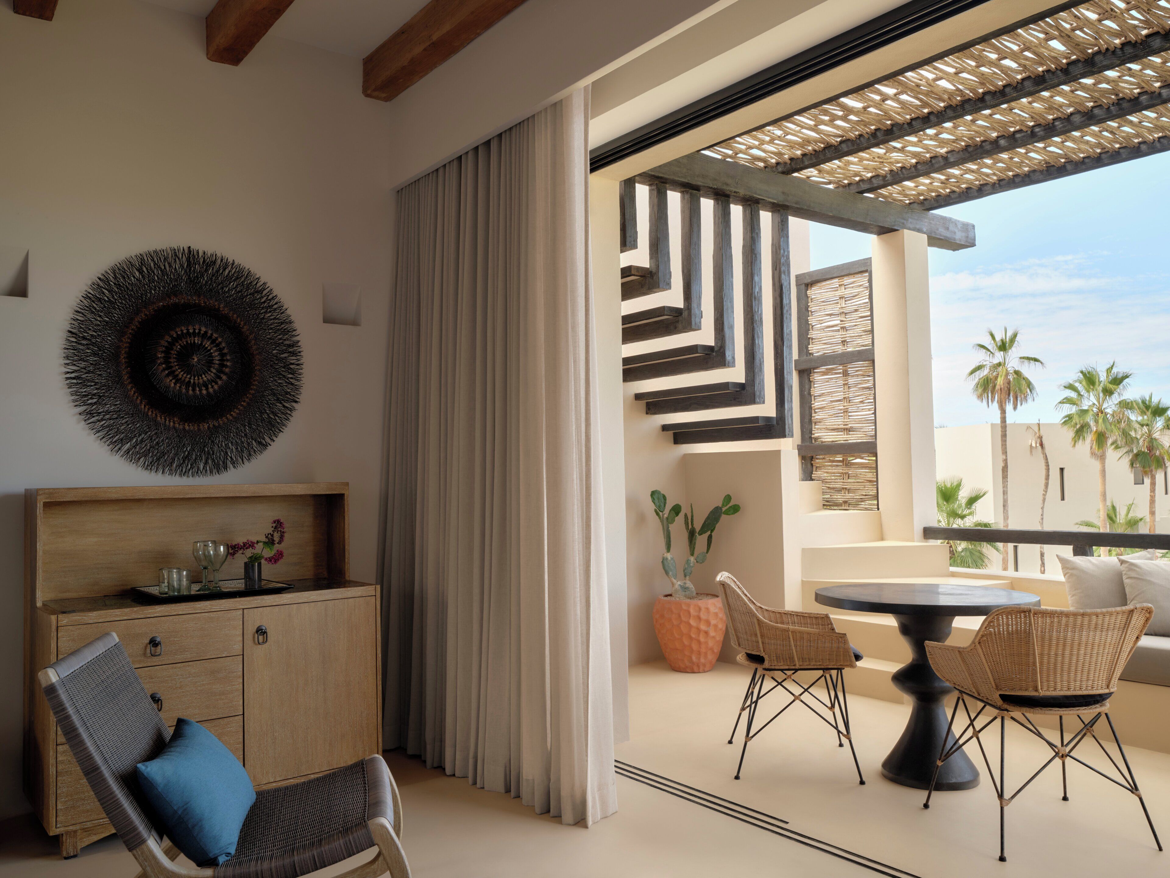 A modern interior with a wooden cabinet, wicker chairs, a round table, and a view of palm trees through a window.