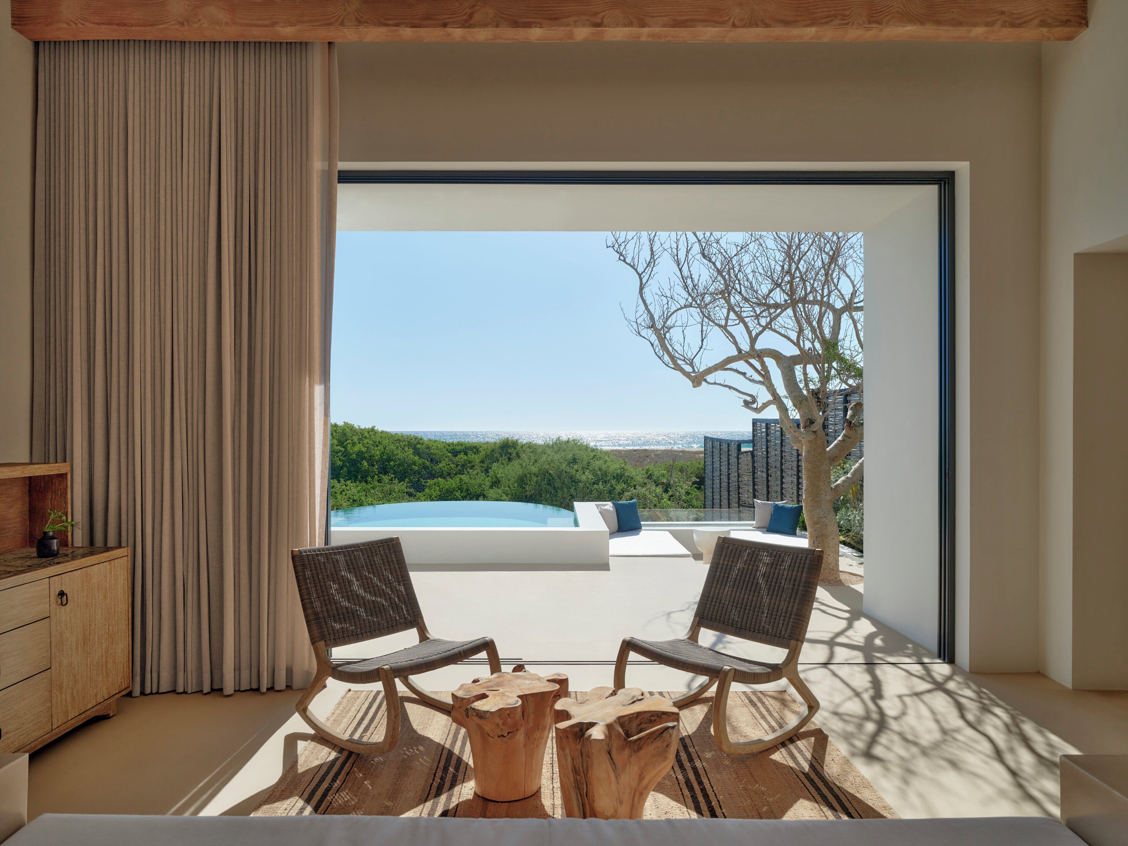 A modern room with a large window, two chairs, a wooden table, and a view of a swimming pool and greenery.