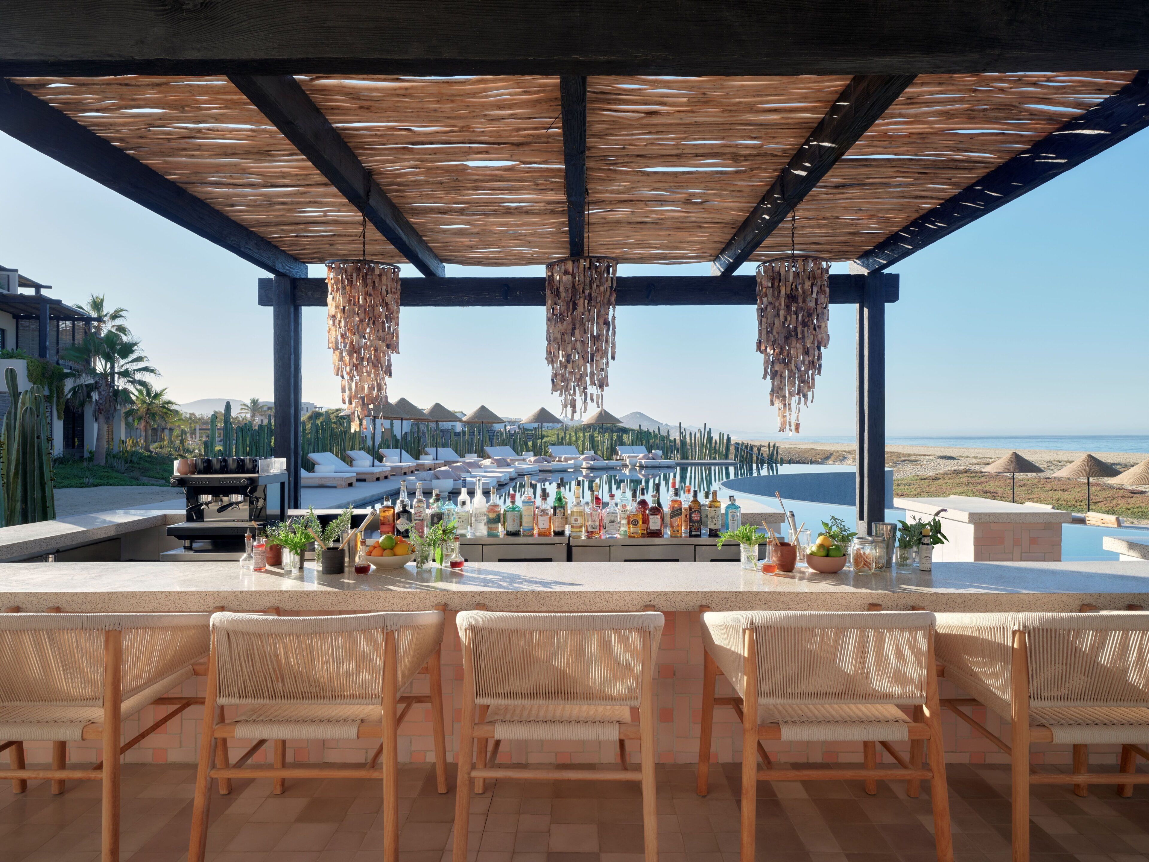 Bar al aire libre techado con asientos, piscina y vista a la playa y palmeras.