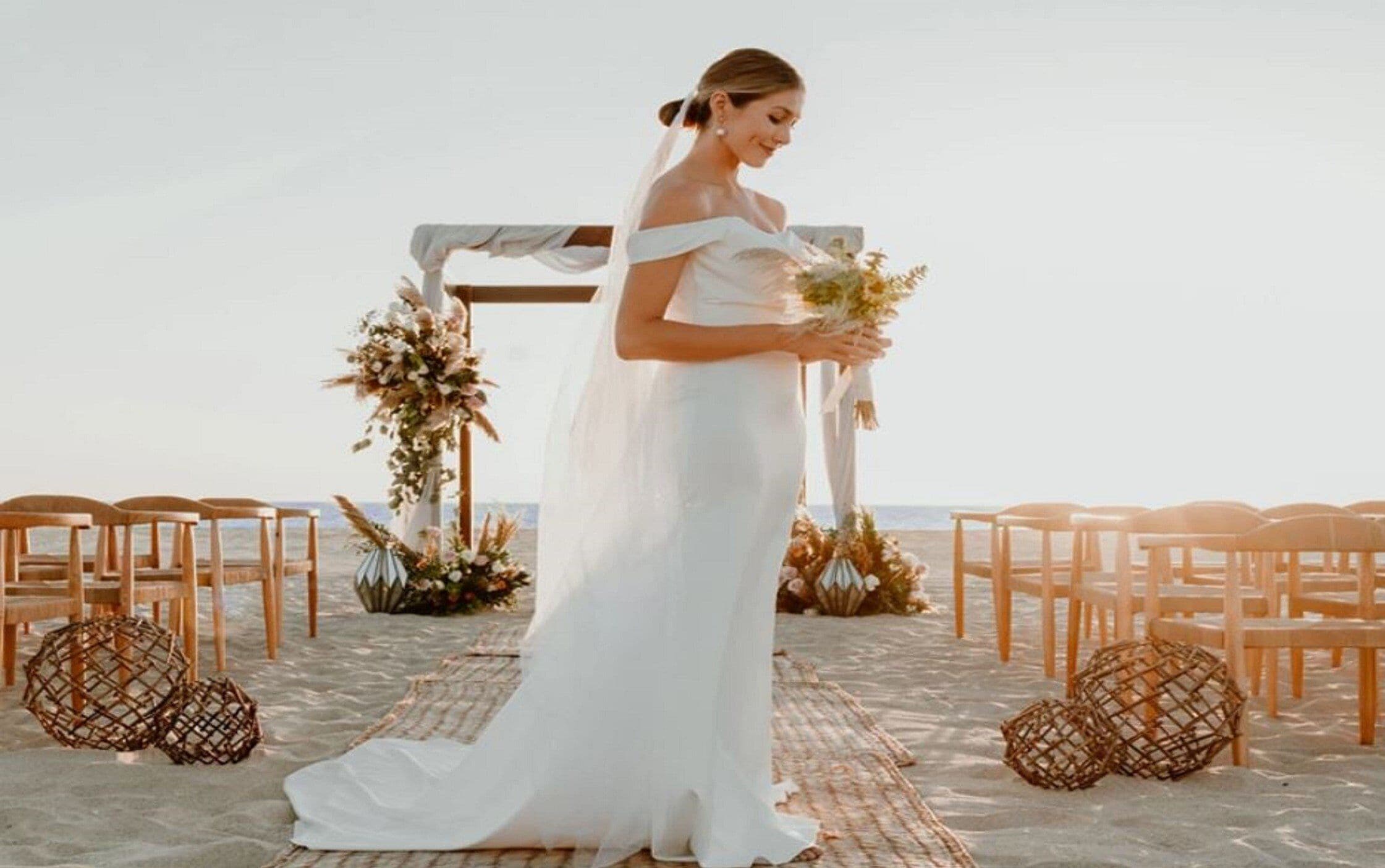 En strandbryllupsopsætning med en brud i hvid kjole, træstole og blomsterpynt.