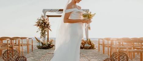 En strandbryllupsopsætning med en brud i hvid kjole, træstole og blomsterpynt.