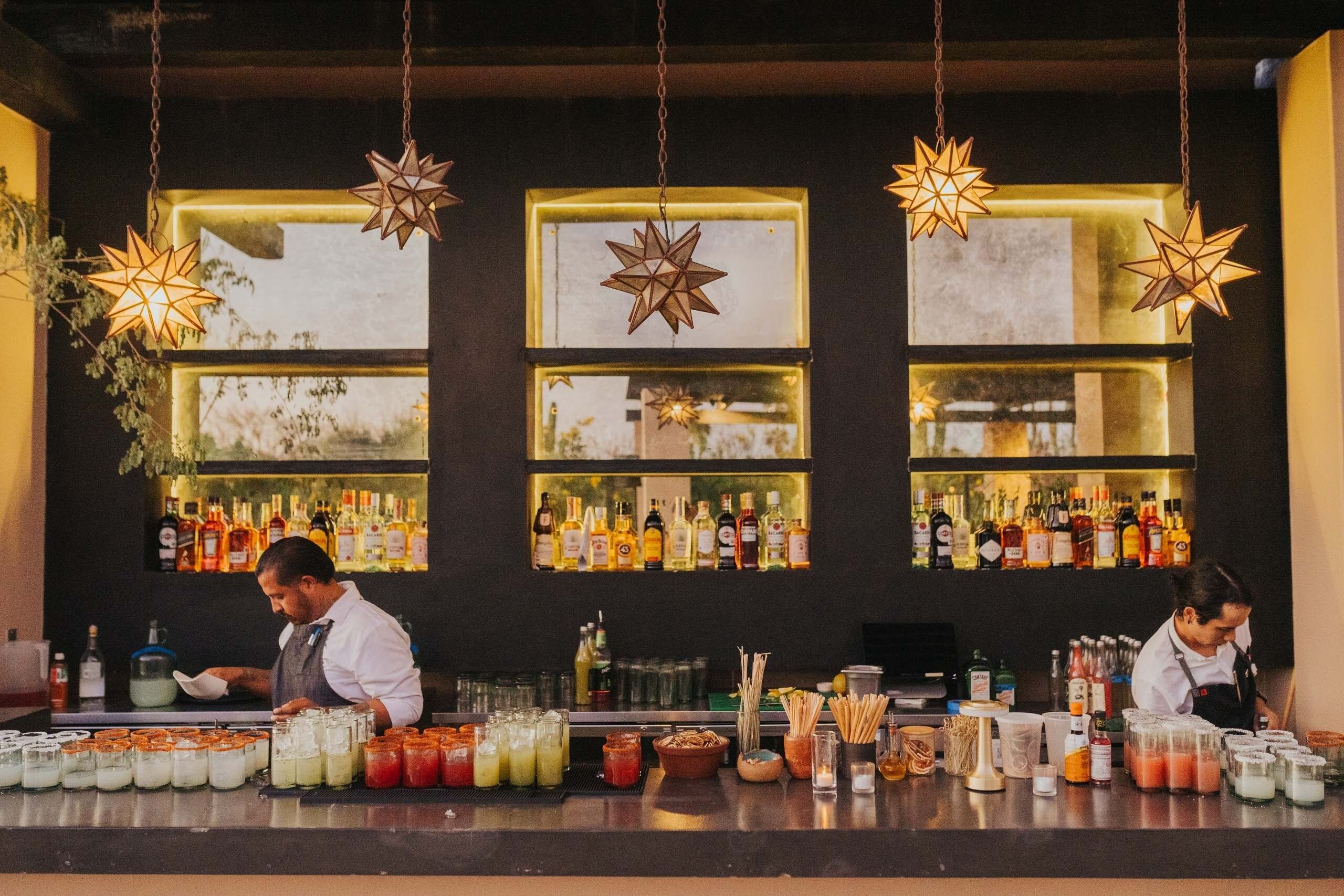 Un bar con múltiples estantes llenos de botellas de licor, un barman preparando bebidas e iluminación decorativa con luces en forma de estrella.
