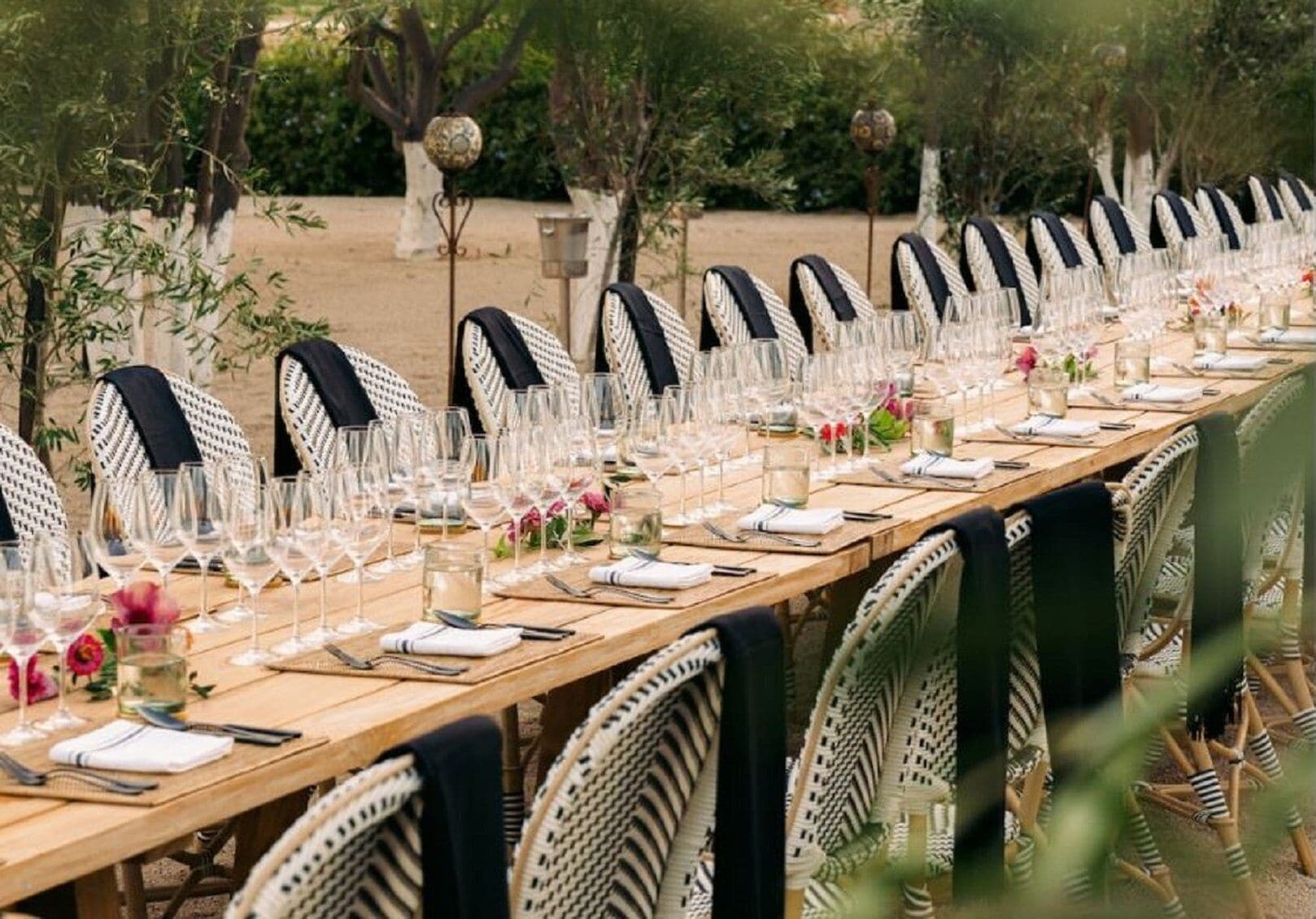 Une longue table à manger installée à l’extérieur, avec des chaises en osier, de la vaisselle transparente et des centres de table floraux.