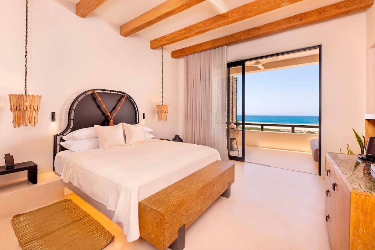A bedroom with a large bed, wooden beams, a nightstand, a bedside table, and a view of the beach.