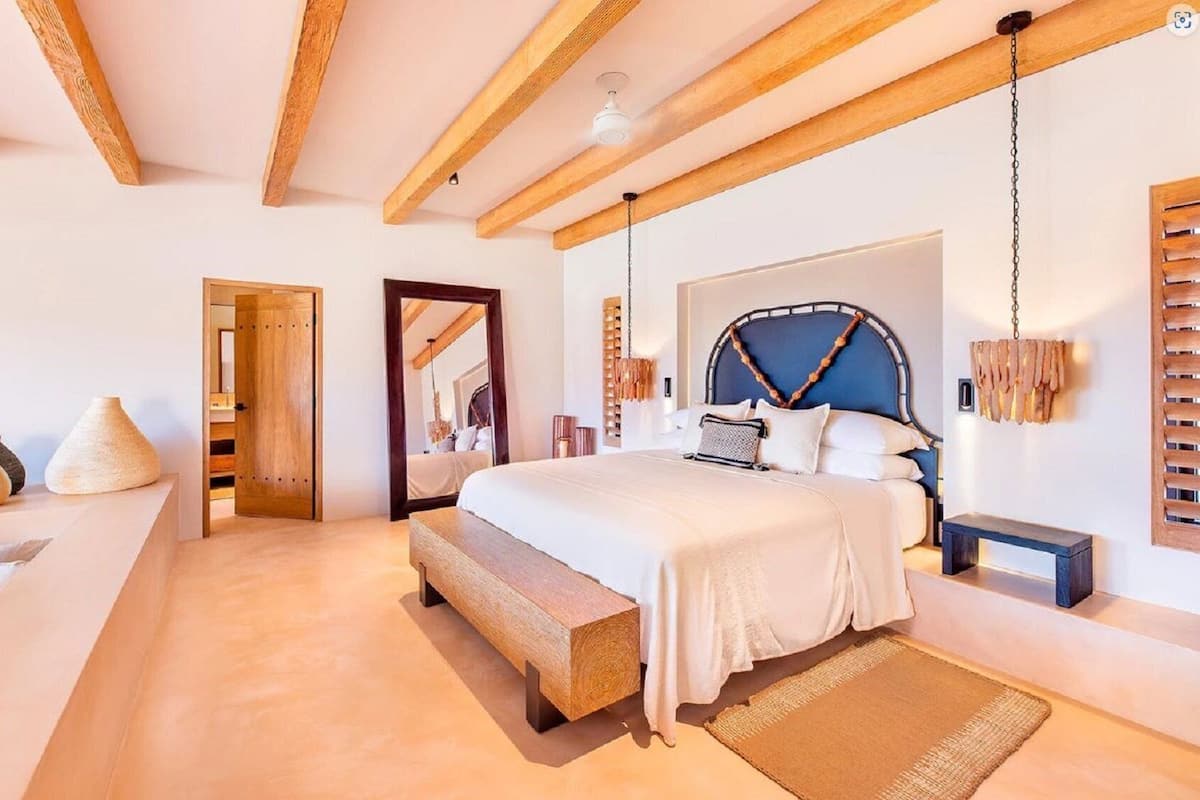 A bedroom with a bed, wooden beams, a hanging light fixture, and a wooden bench.
