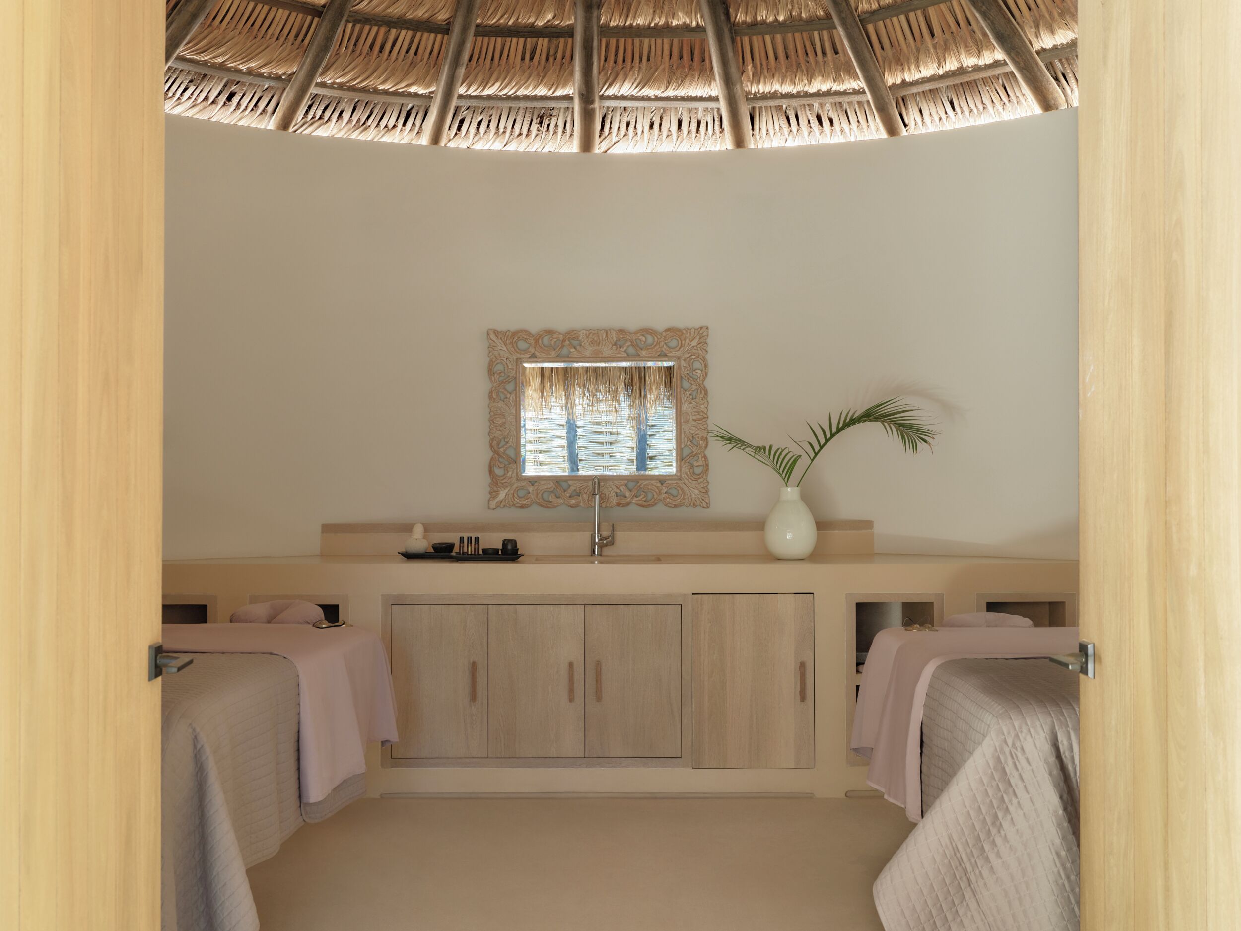 Un baño con techo de paja, un lavamanos con espejo decorativo, un florero con planta y armarios de madera.