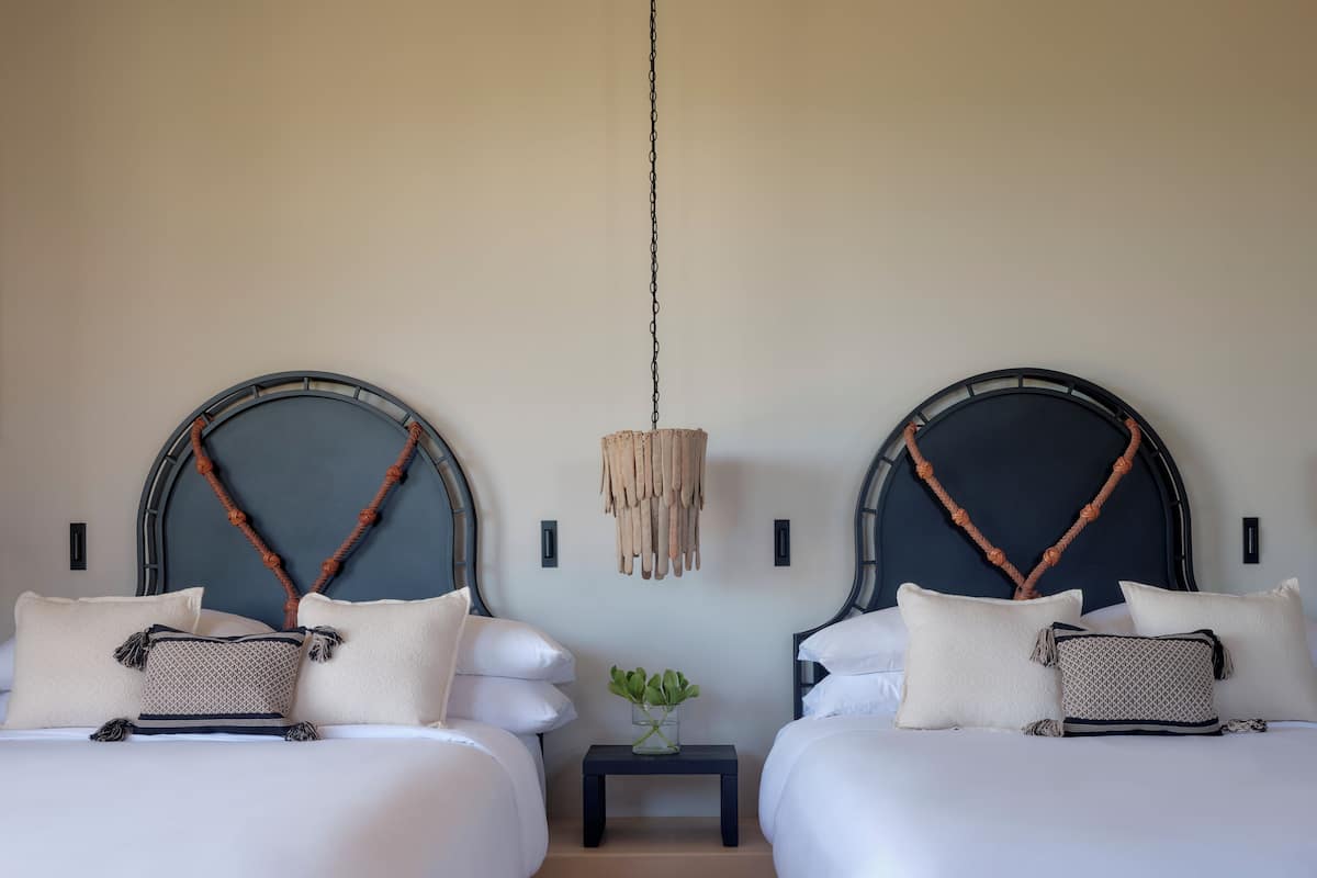 A hotel room with two beds, each with a unique headboard design, a small bedside table with a plant, and a hanging light fixture.