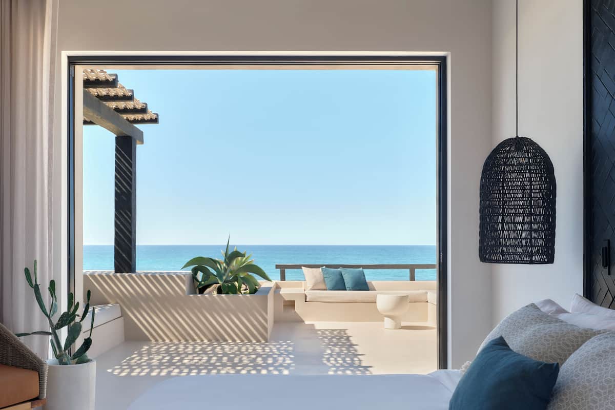 A modern hotel room with a large bed, a view of the sea, a balcony with a sofa, and a cactus plant.