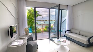 Un salon moderne avec un canapé, une table basse et un téléviseur, donnant sur une terrasse offrant une vue sur la mer et les palmiers.