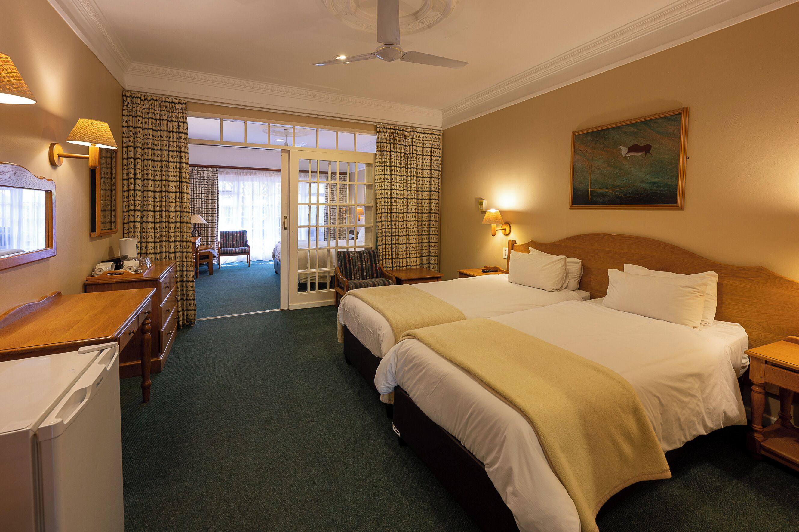 A hotel room with two beds, a desk, a chair, a wardrobe, a ceiling fan, and a painting on the wall.