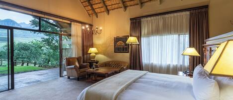 Une chambre d’hôtel avec un grand lit, un canapé, un fauteuil et une table. Il y a deux lampes sur la table et un lustre au plafond. La chambre est dotée de grandes fenêtres avec des rideaux et offre une vue sur l’extérieur.
