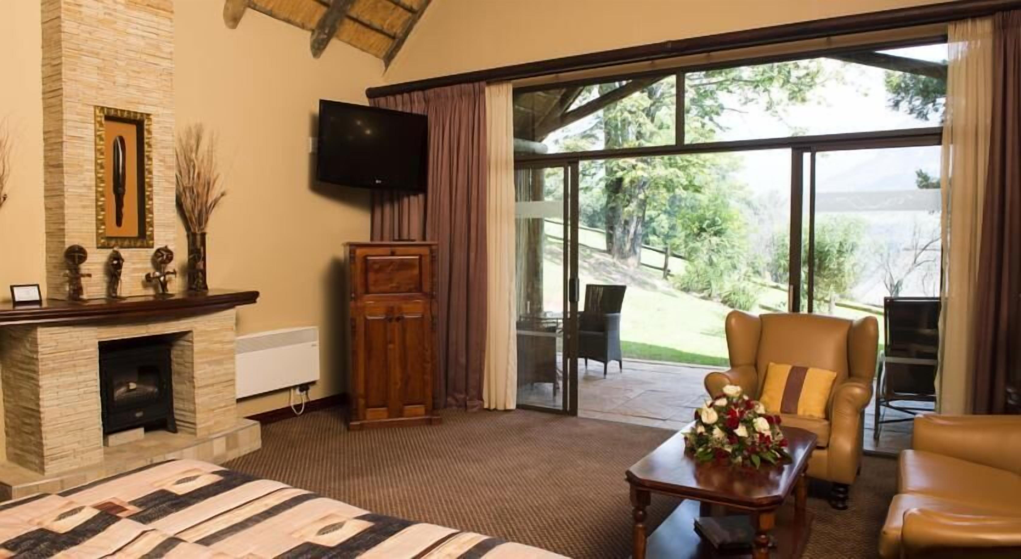 A spacious room with a large bed, a fireplace, a television, and a view of a garden through large windows.
