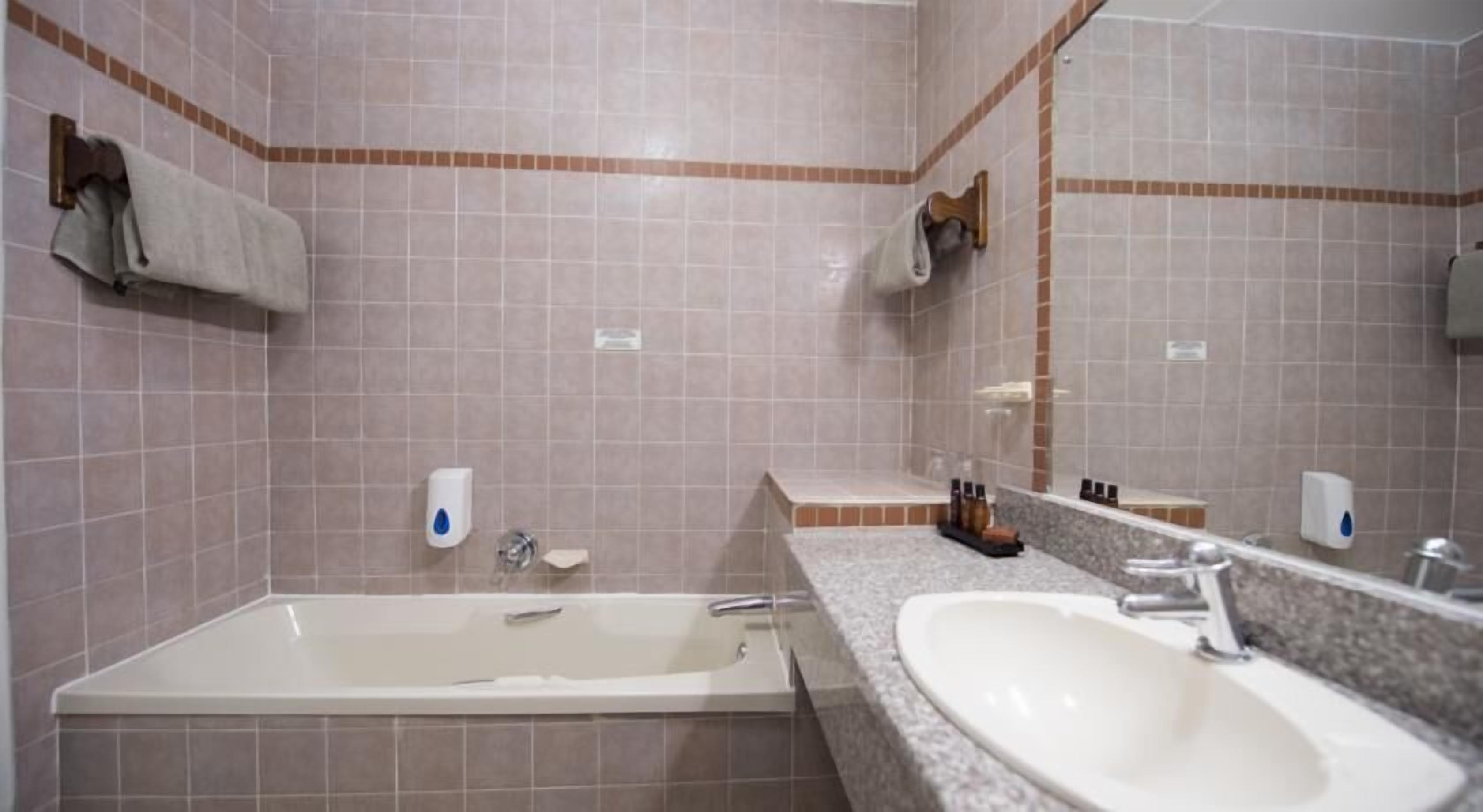 A bathroom with a bathtub, a sink, and a mirror. There are towels hanging on hooks and soap dispensers on the wall.