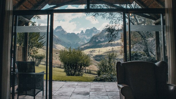 A room with a large glass door leading to a scenic mountain view. Inside, there are two chairs and a stone floor.