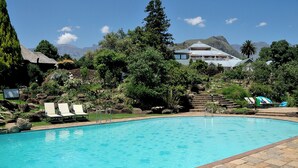 A swimming pool with lounge chairs, surrounded by greenery and mountains in the background.