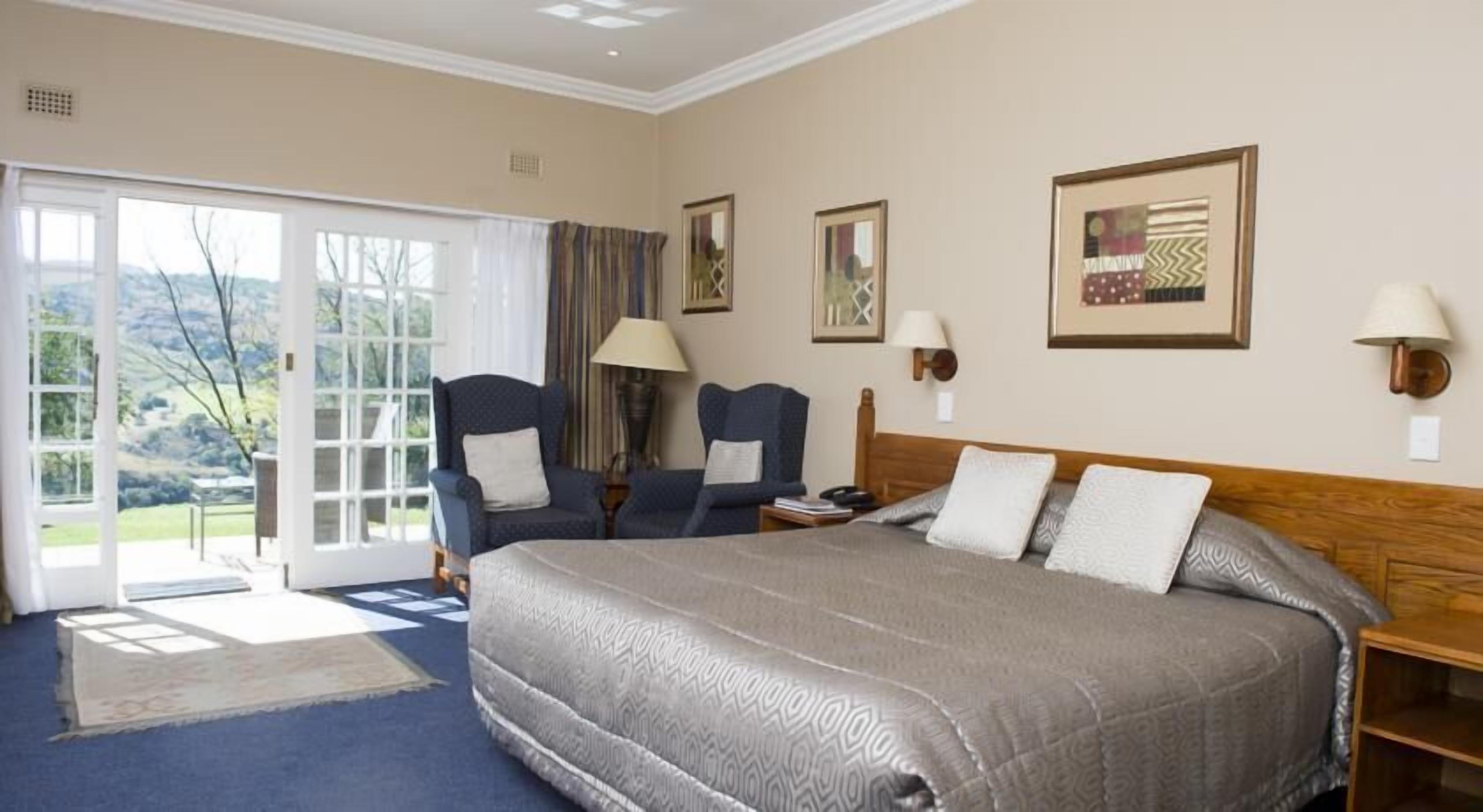 A hotel room with a large bed, two armchairs, a desk, and a view through the glass doors.