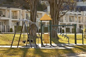 A playground with swings and a slide, surrounded by trees and a building in the background.