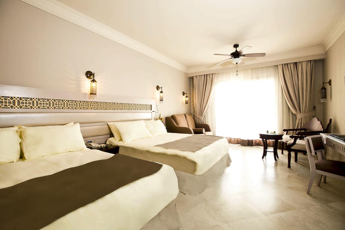 A hotel room with two beds, a desk, a chair, a ceiling fan, and a large window with curtains.