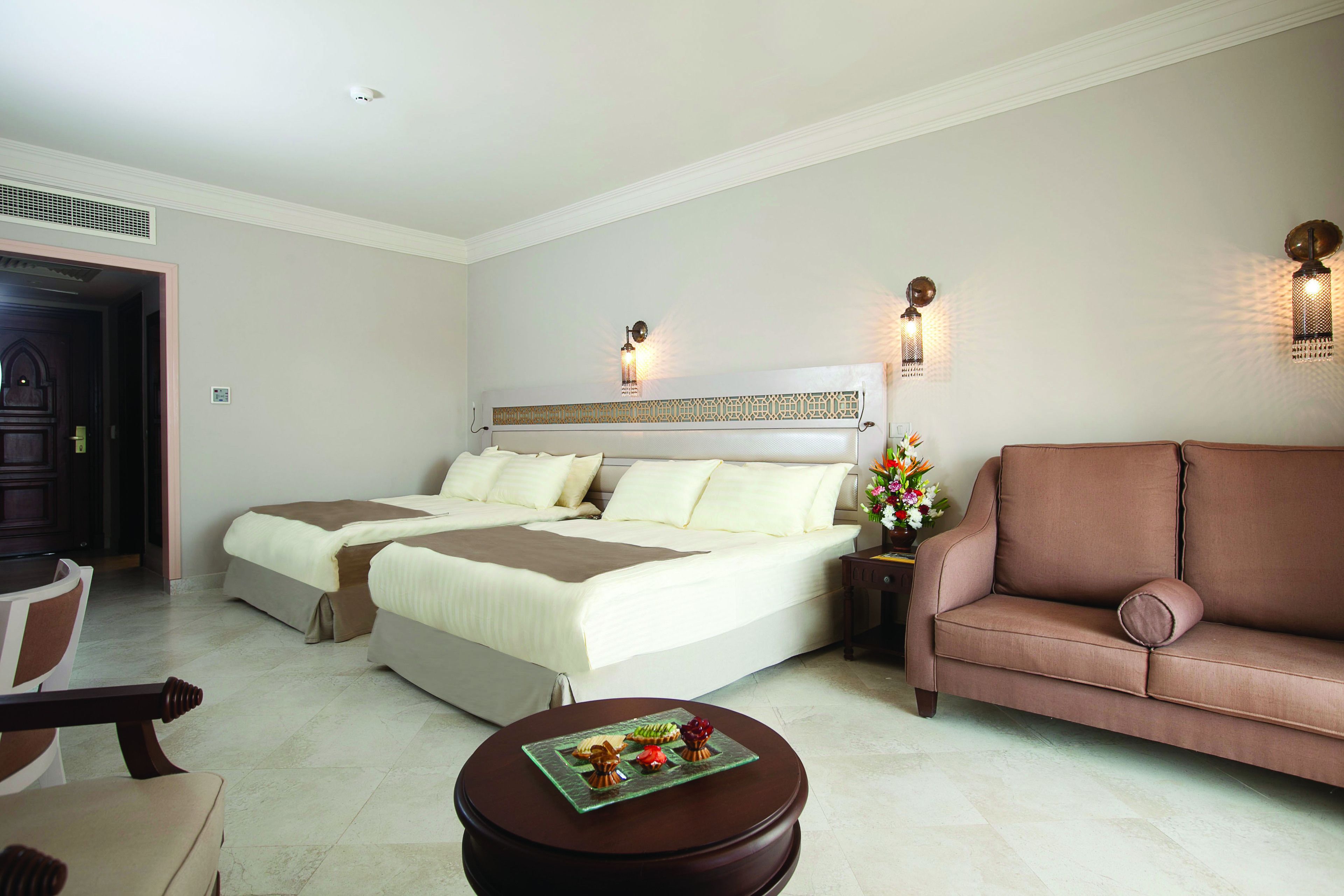 A hotel room with a double bed, a sofa, a small table with decorative items, and wall-mounted lights.