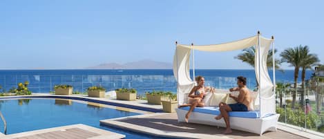 A couple relaxing on a white wicker sofa by a pool with a view of the ocean and mountains.