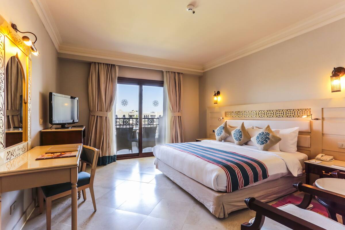 A hotel room with a bed, a desk, a chair, a television, a balcony with curtains, and a view of the city.