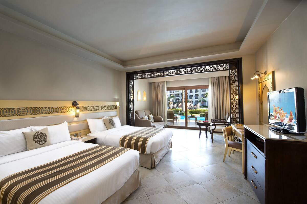 A hotel room with two beds, a TV, a desk, and a view of a pool and building.