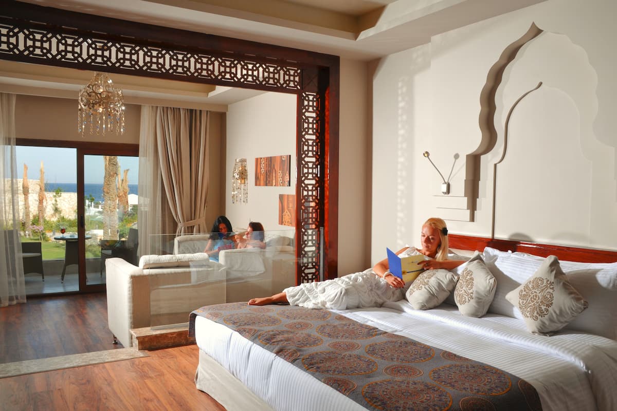 A spacious bedroom with a large bed, a glass door leading to a balcony, and a view of the sea.
