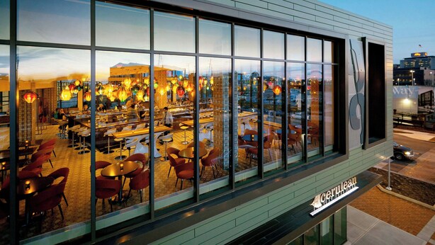 A modern building with large windows reflecting a well-lit interior space, possibly a restaurant or cafe, with visible seating areas and a clear view of the cityscape in the background.