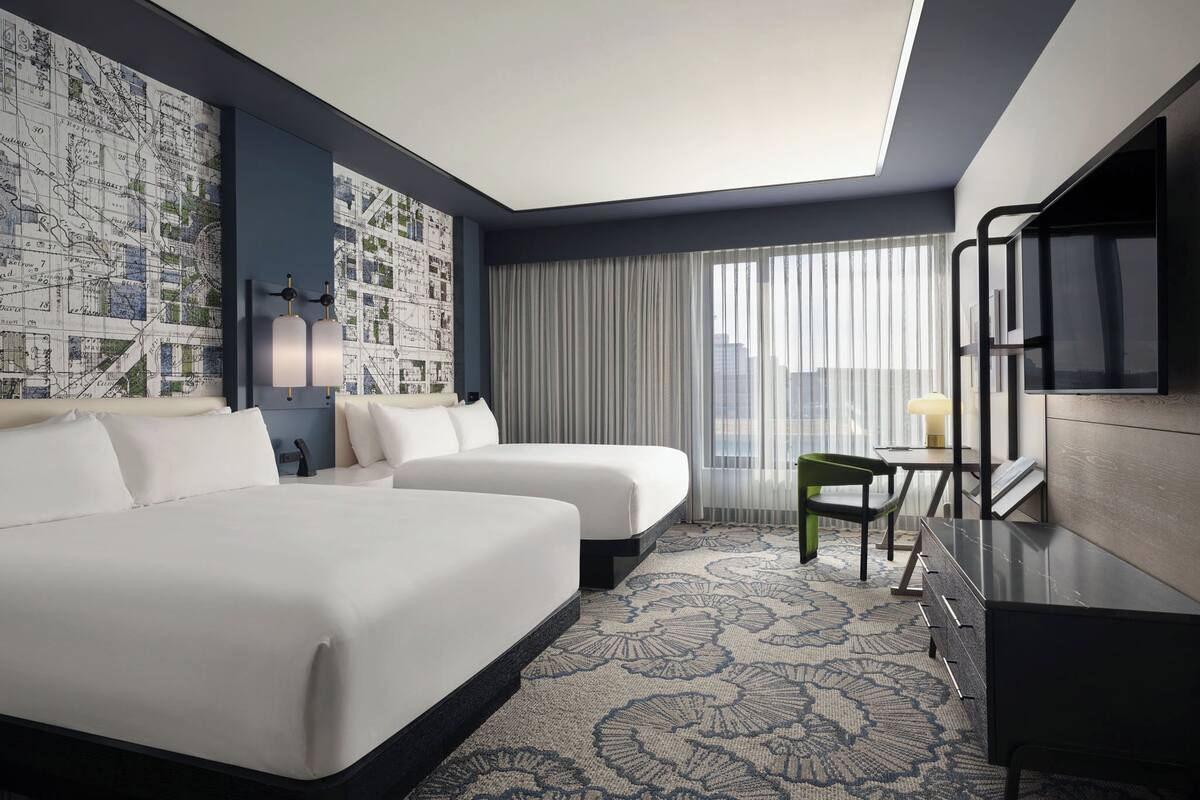 A hotel room with two beds, a desk, a chair, a TV, and a large window with curtains.