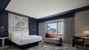 A hotel room with a large bed, a sofa, a desk, and a view of a cityscape through the window.
