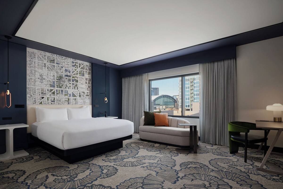 A hotel room with a large bed, a sofa, a desk, and a view of a cityscape through the window.