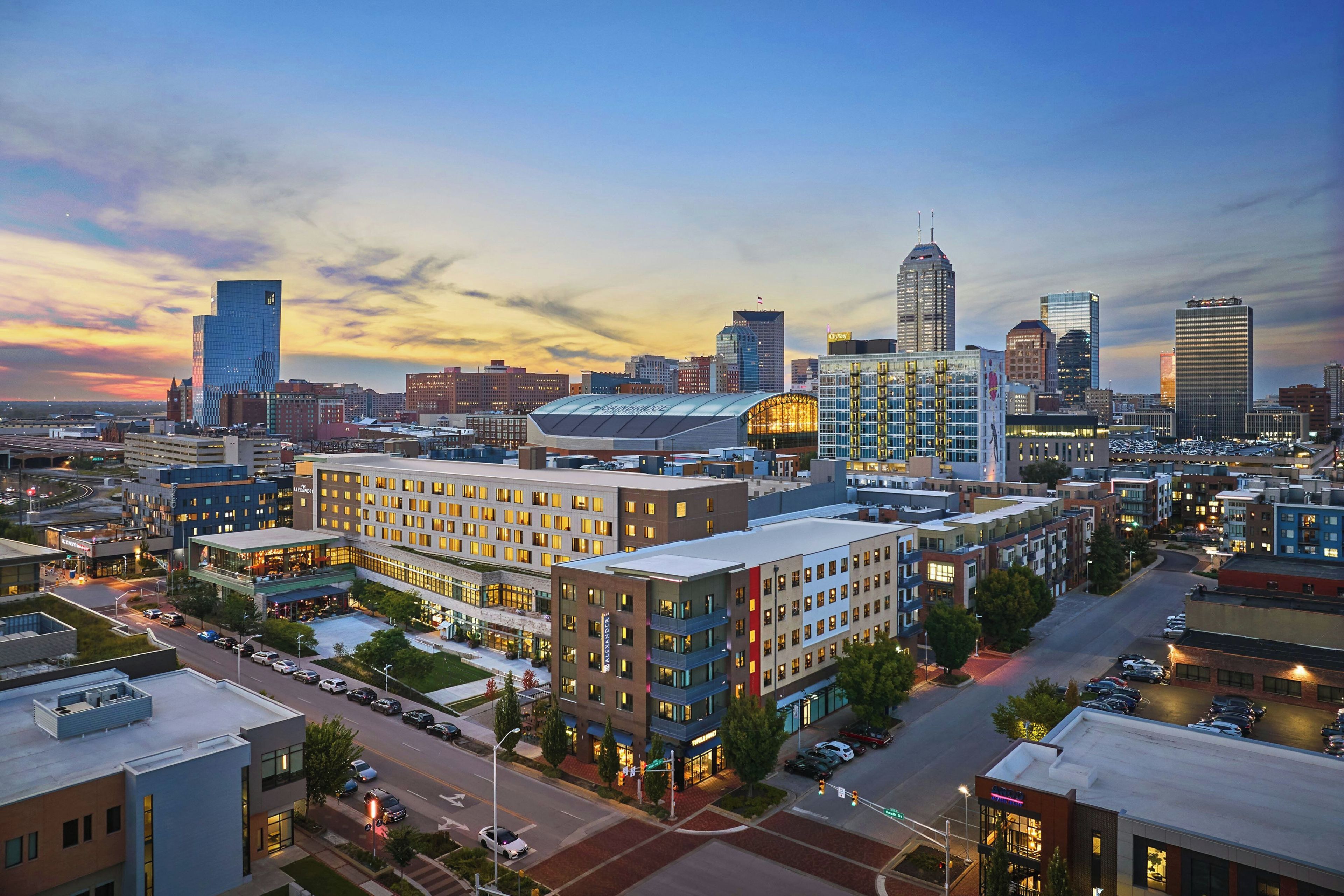 A cityscape with modern buildings, a prominent glass skyscraper, and a well-lit hotel with a large parking lot.