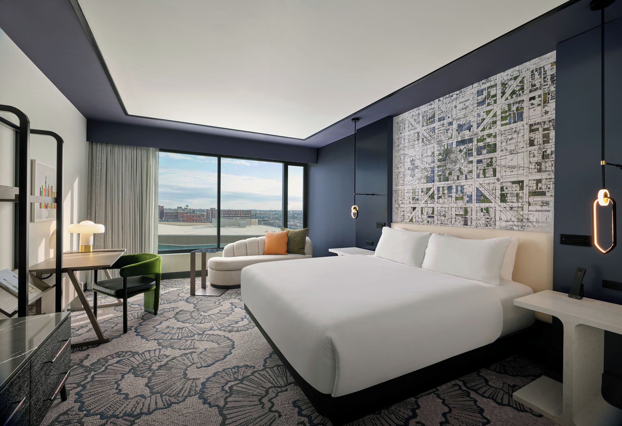 A modern hotel room with a large bed, a desk with a chair, a sofa, and a cityscape view through the window.