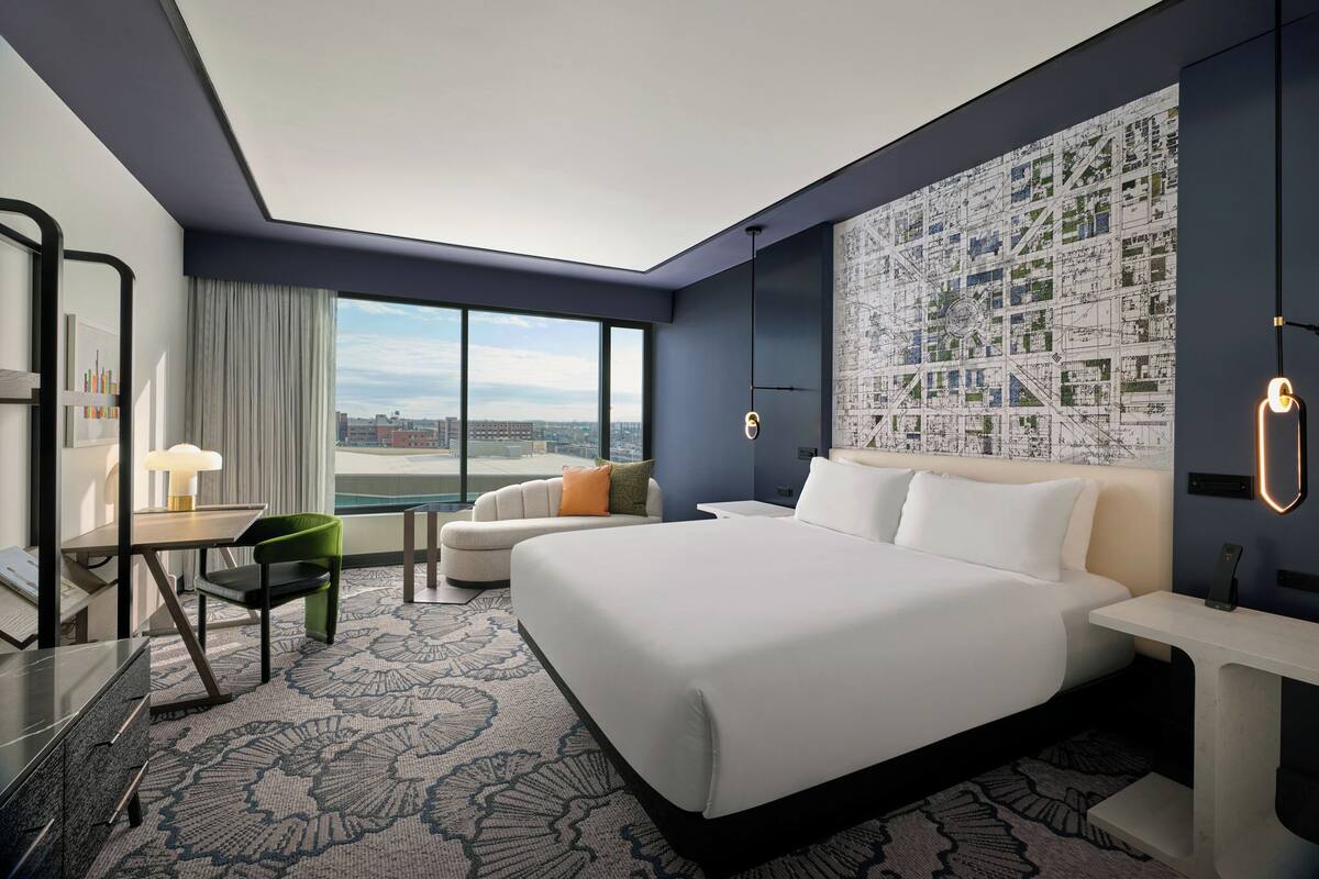 A modern hotel room with a large bed, a desk with a chair, a sofa, and a cityscape view through the window.